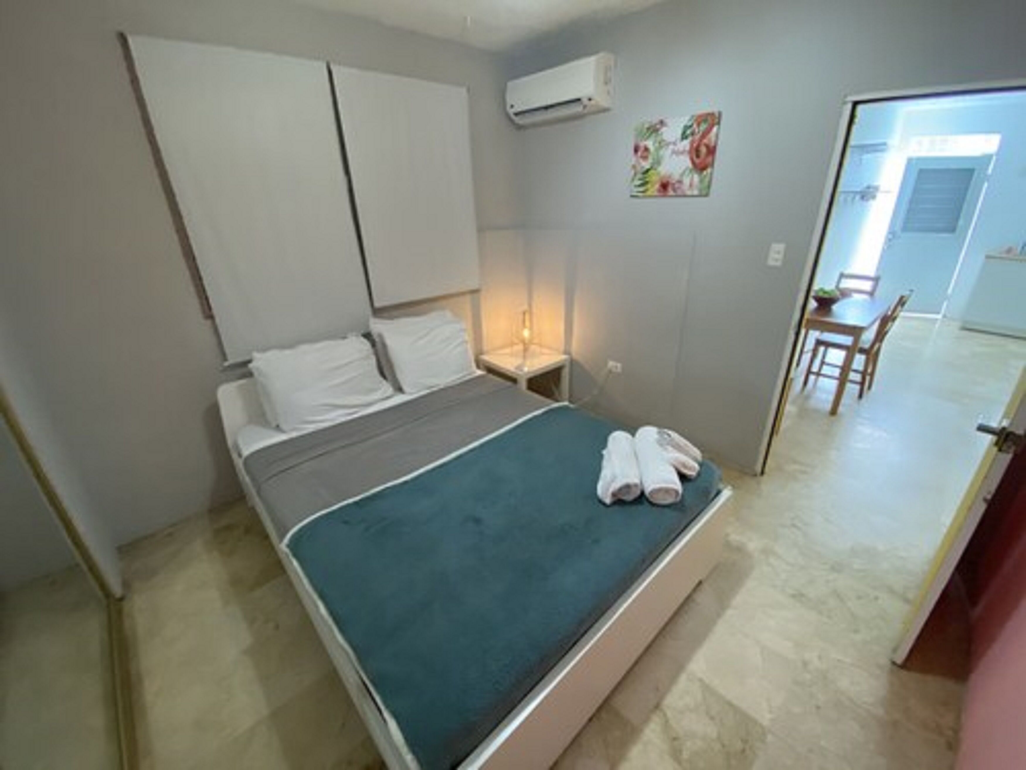 A bedroom with a bed, a nightstand, an air conditioning unit, a painting, and a dining area visible through an open door.