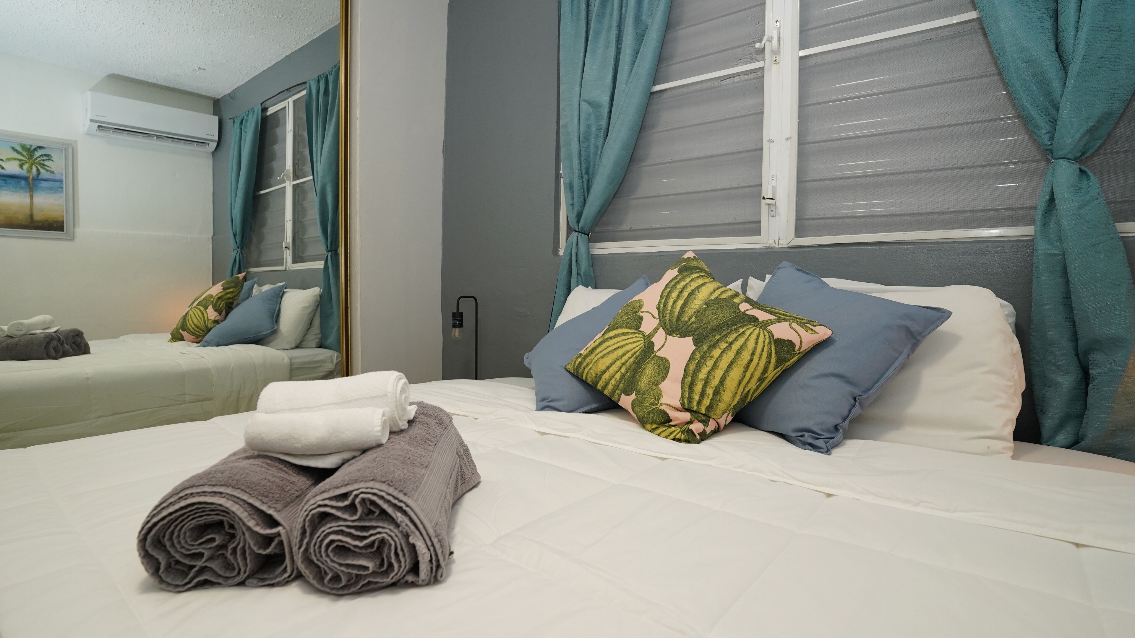 Een opgemaakt bed met opgerolde handdoeken, een raam met gordijnen en een schilderij aan de muur.