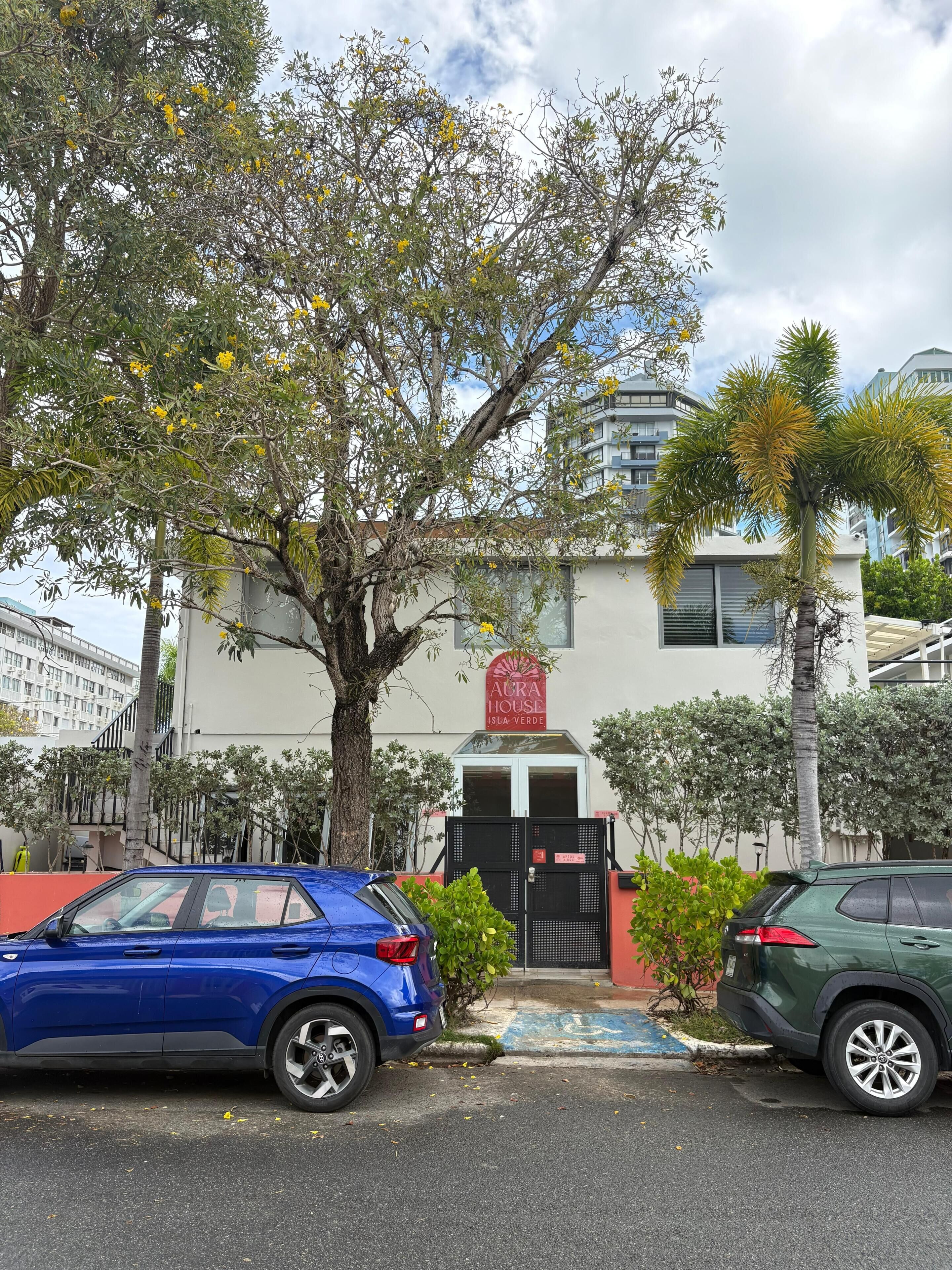 Un bâtiment avec un portail noir, une enseigne indiquant « AURA HOUSE ISLA VERDE », et deux voitures garées.
