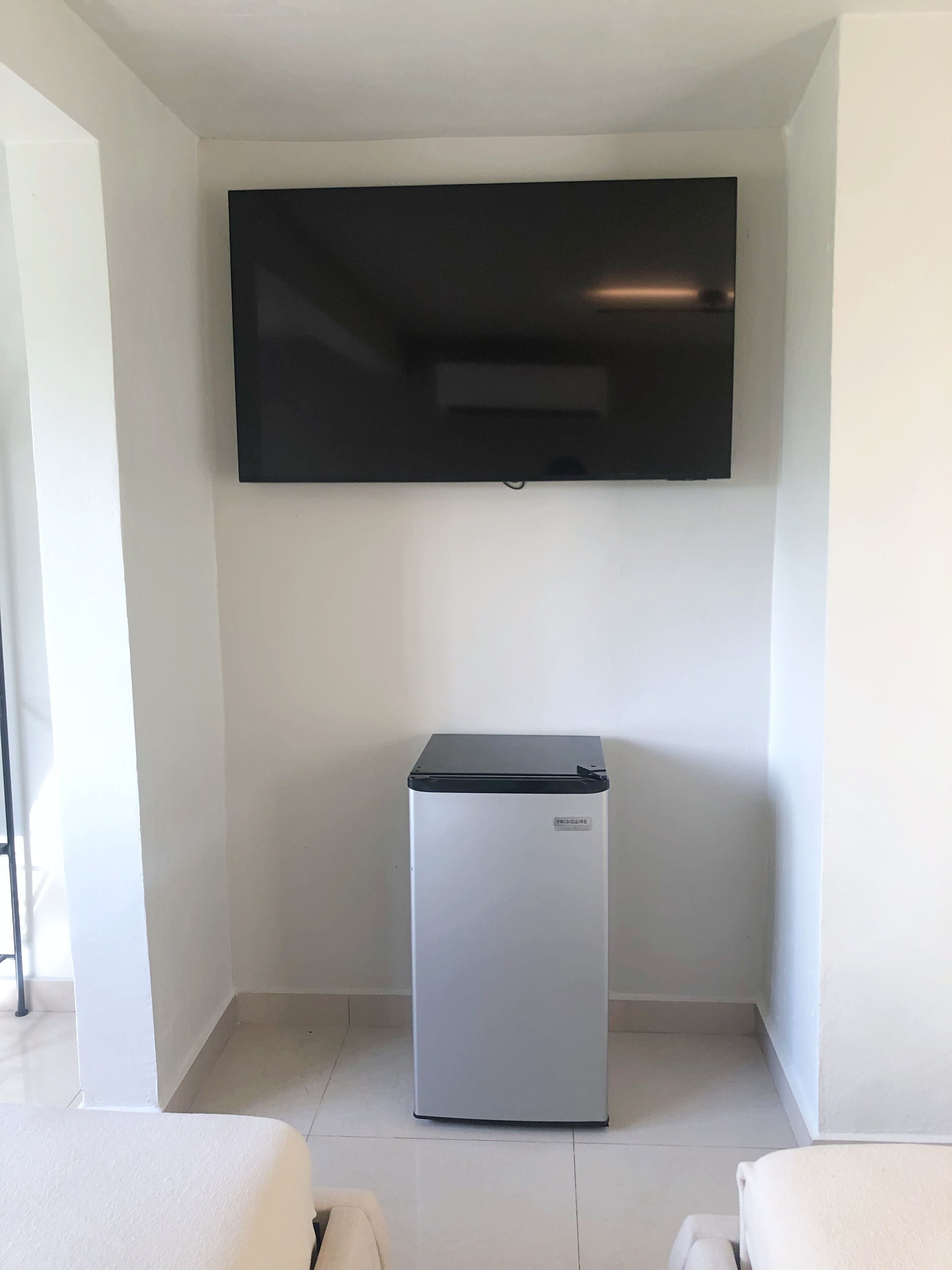 A compact room with a flat-screen TV mounted on the wall, a small refrigerator, and a white chair.