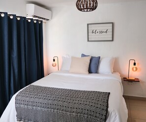 A neatly made bed with a patterned throw, two bedside lamps, a wall-mounted air conditioner, and a framed sign saying "blessed".