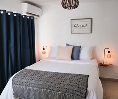 Una cama bien hecha con una manta estampada, dos lámparas de noche, un aire acondicionado de pared y un cartel enmarcado que dice "blessed".