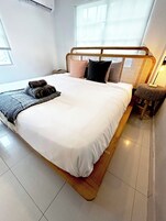 Una cama bien tendida con cabecera de madera, una mesita de noche y una silla pequeña.