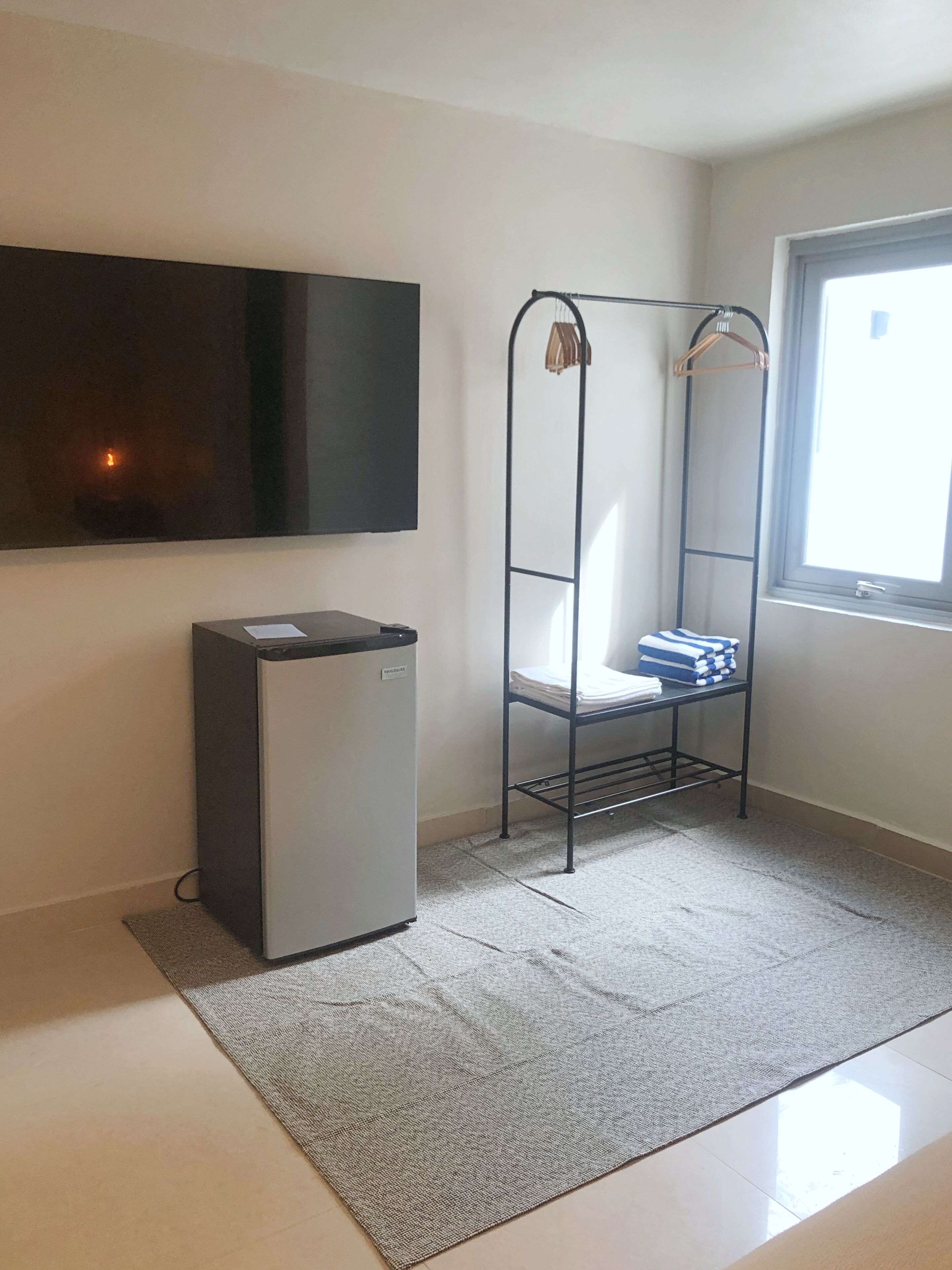 Une petite pièce équipée d’un téléviseur à écran plat fixé au mur, d’un réfrigérateur compact, d’une étagère en métal où sont pliés des serviettes et d’une fenêtre laissant entrer la lumière naturelle.