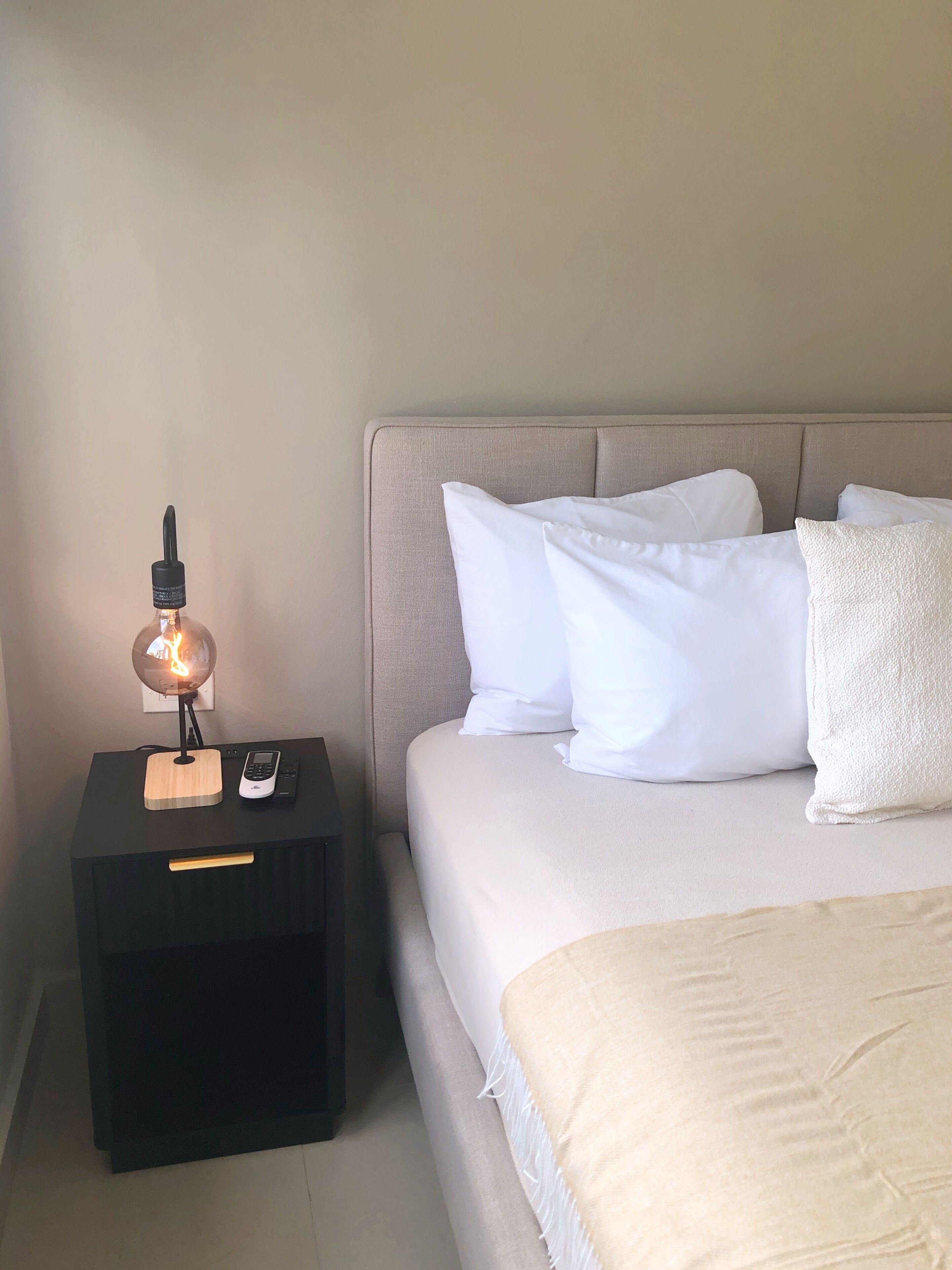 Una cama bien hecha con ropa blanca y almohadas, una mesita de noche con lámpara y un control remoto.