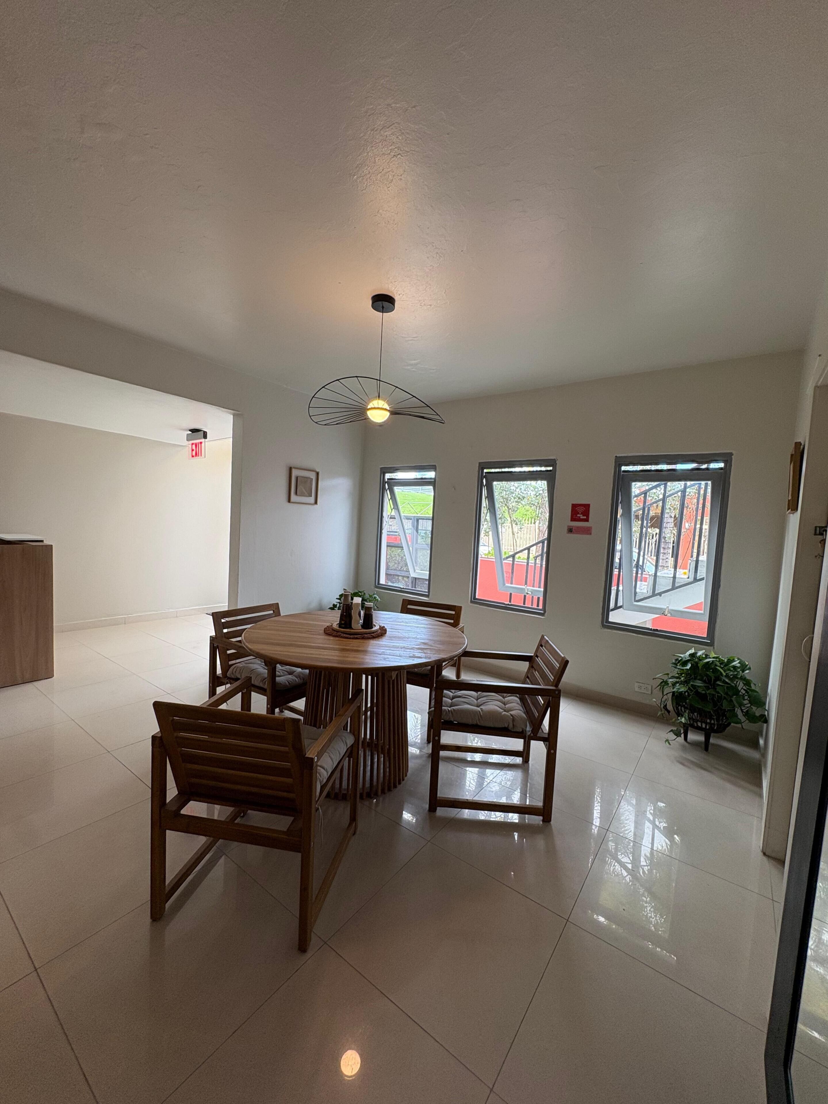 A spacious room with a round wooden table surrounded by chairs, large windows, and a ceiling light fixture.