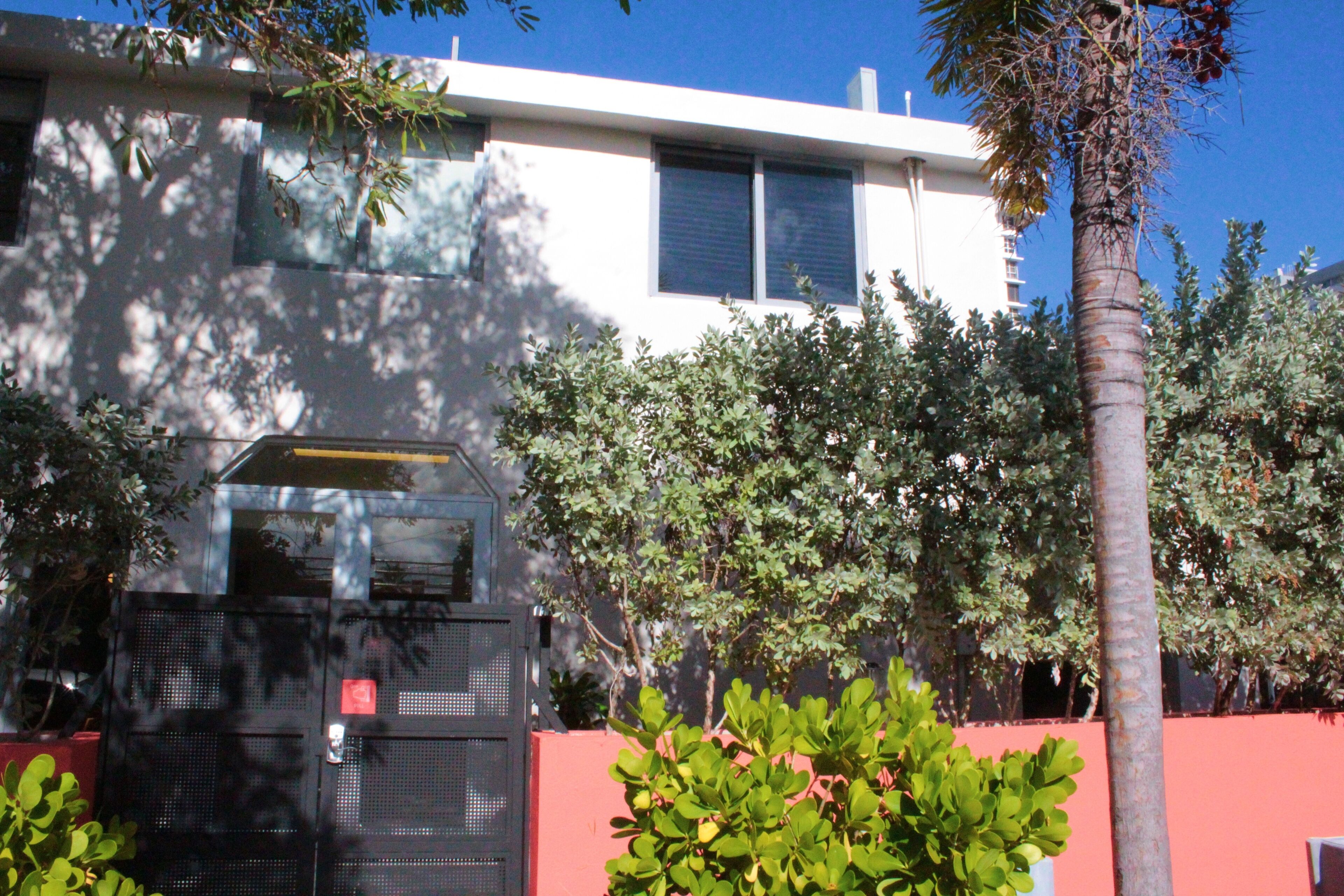 Un edificio moderno con entrada protegida por una reja, rodeado de abundante vegetación y una alta palmera.