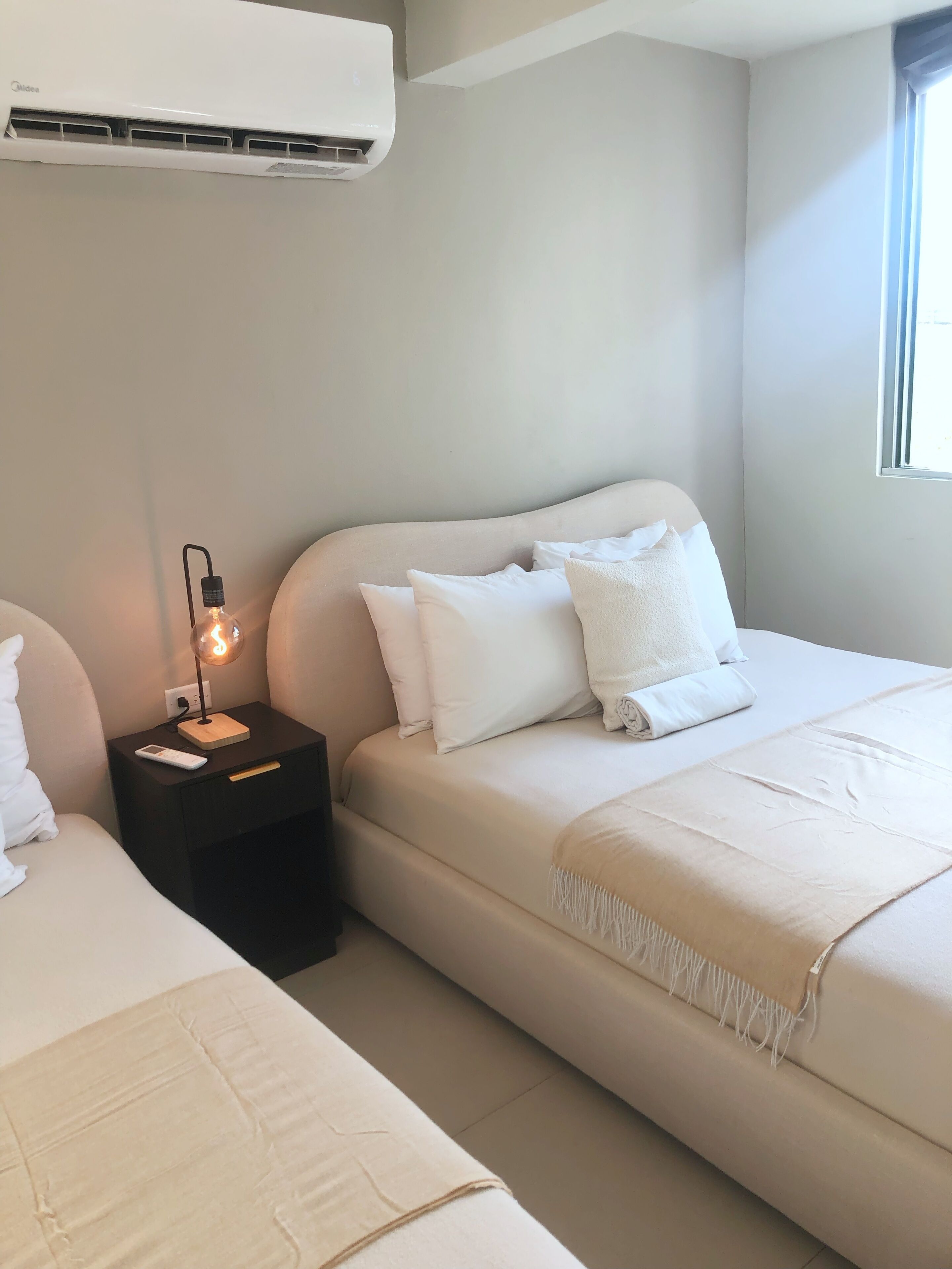 A hotel room with a bed, bedside table, lamp, and an air conditioning unit.