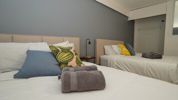 Habitación de hotel con dos camas, cada una con ropa de cama blanca y varias almohadas. Hay una mesita de noche pequeña con una lámpara, y toallas dobladas con cuidado sobre las camas.