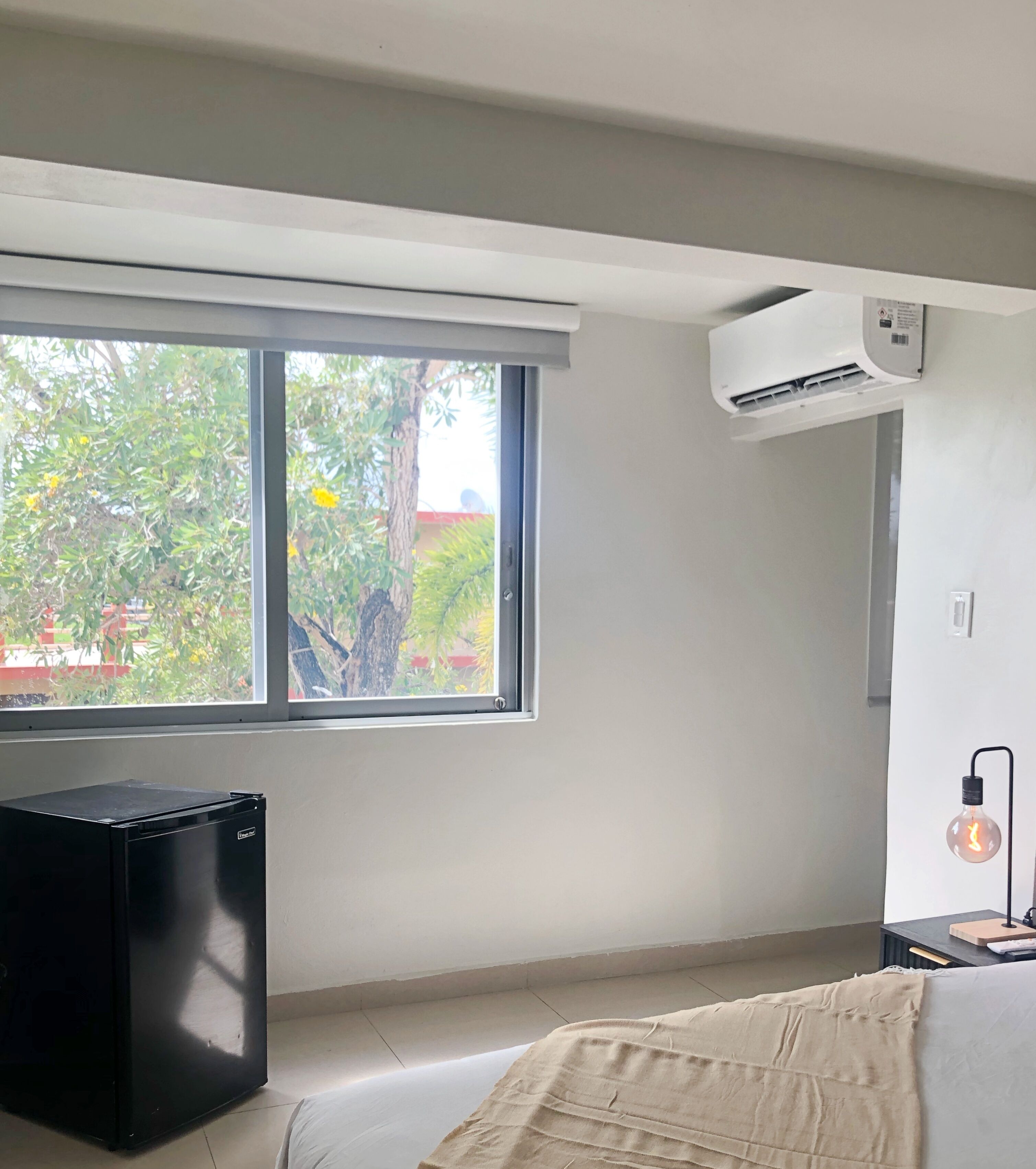 A bedroom with a large window, an air conditioning unit, a black appliance, and a bedside lamp.