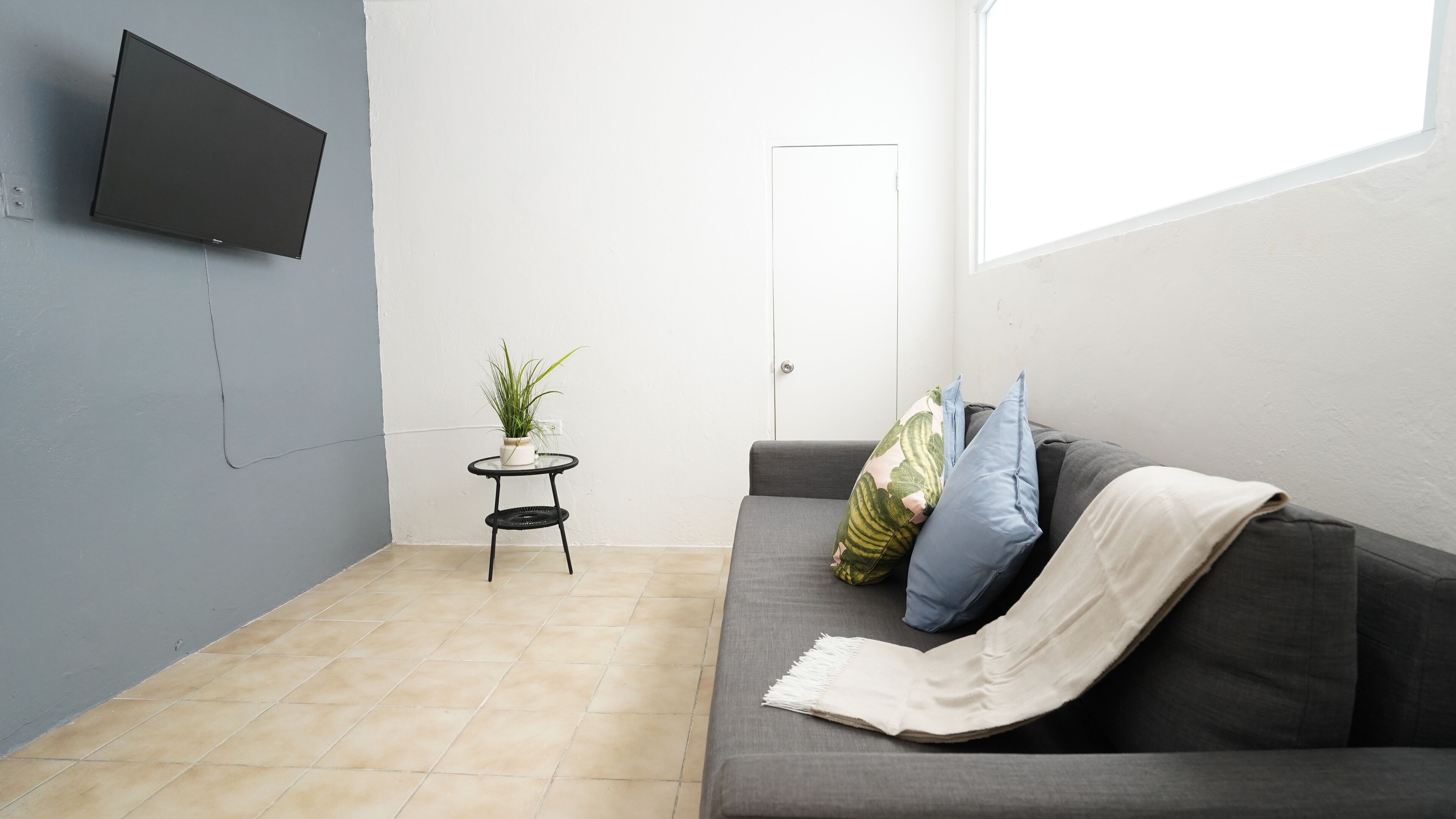 A living room with a grey sofa, a small round table with a plant, and a flat-screen TV mounted on the wall.