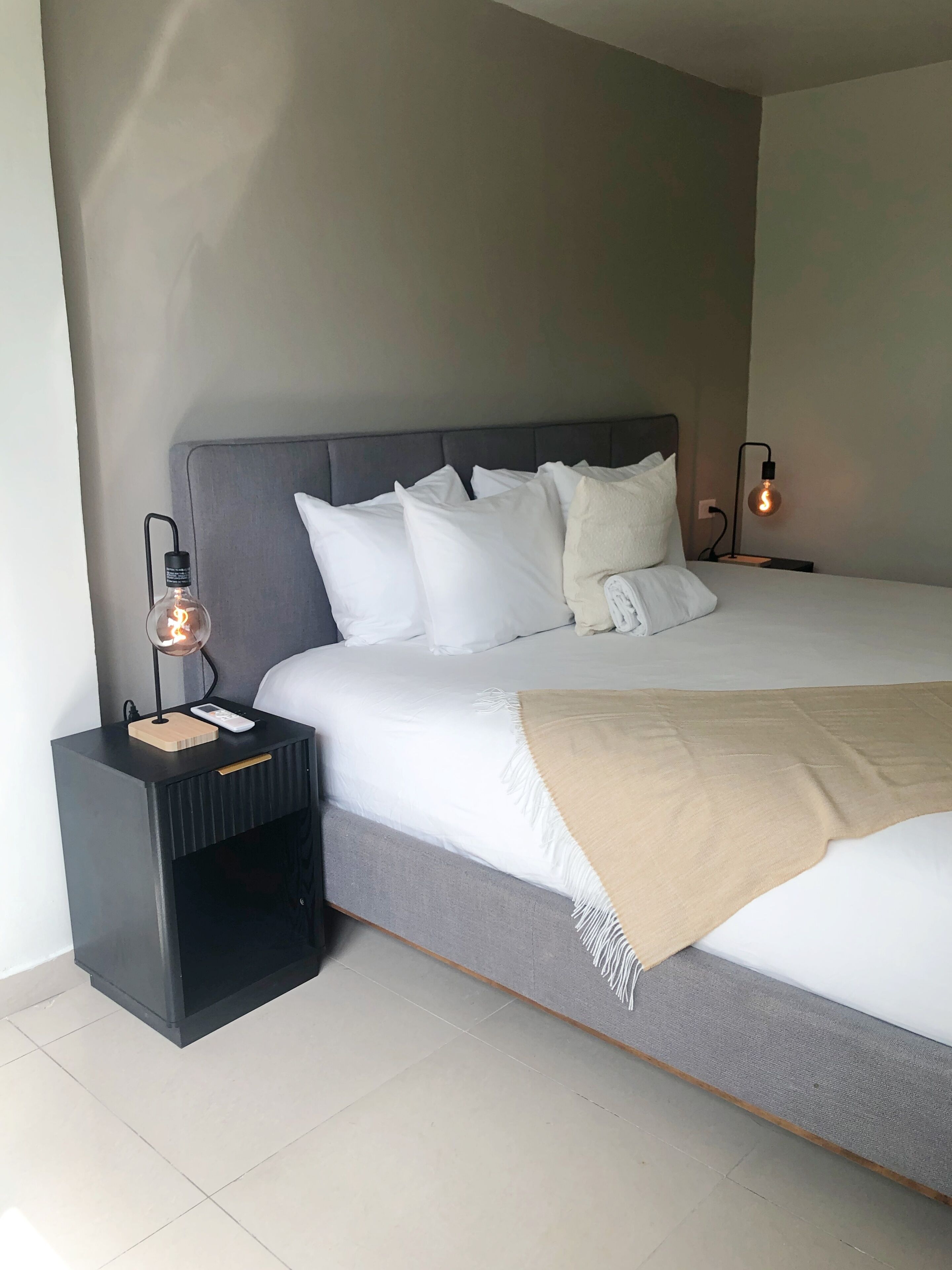 A modern bedroom with a large bed, a bedside table with a lamp, and a nightstand with a phone and charger.