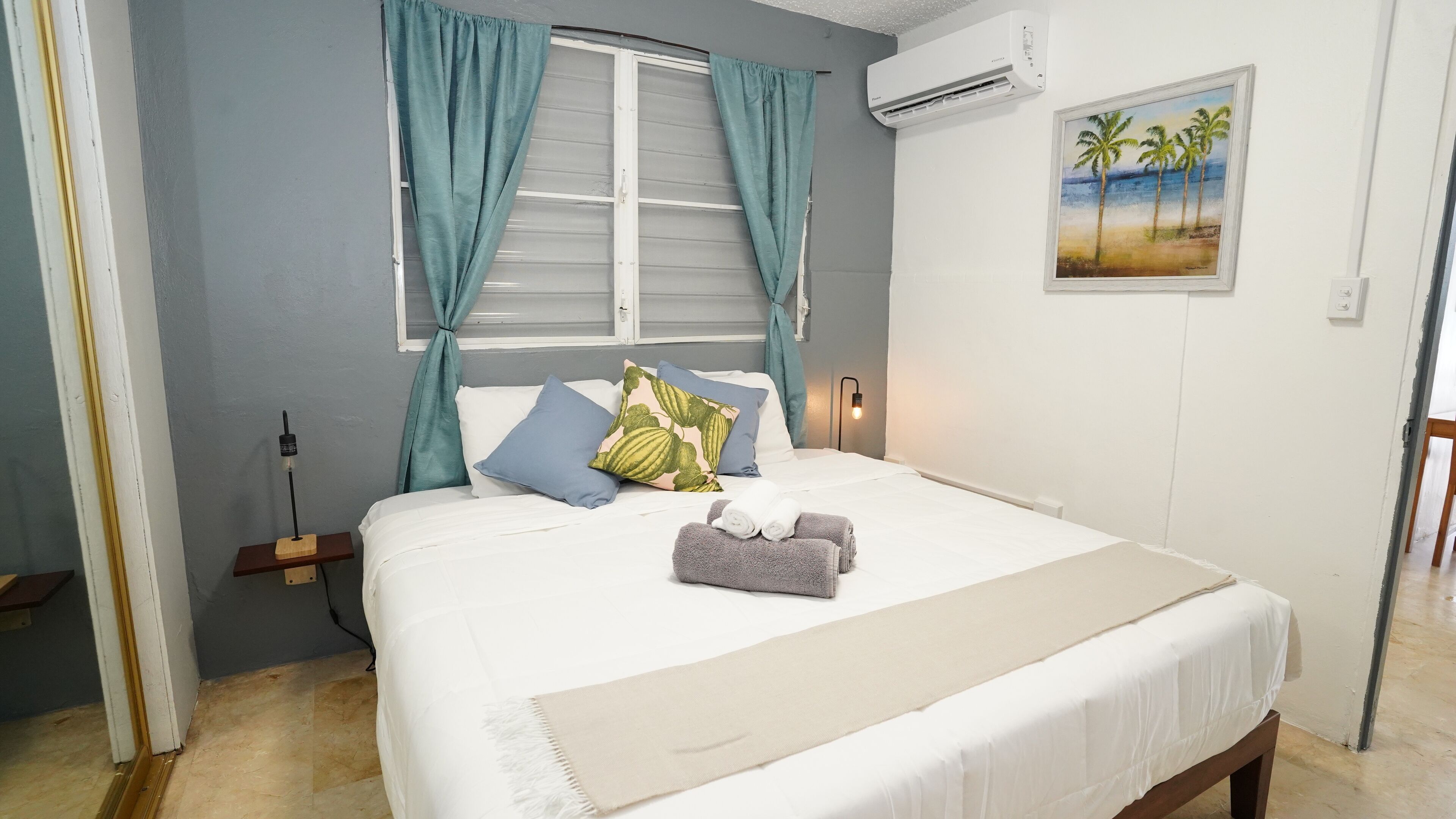 Una cama bien hecha con sábanas blancas, cortinas color turquesa y un cuadro de palmeras en la pared.