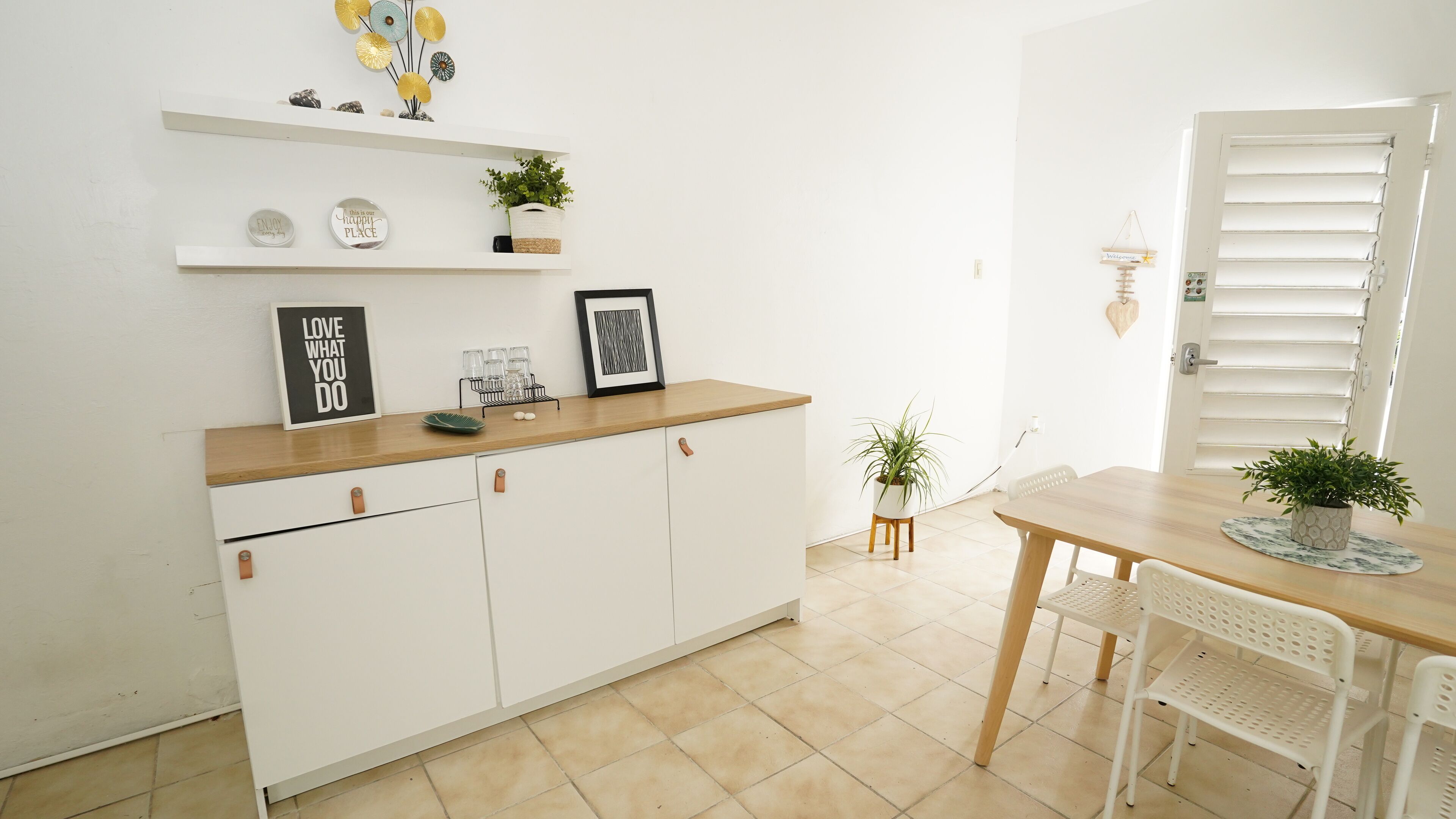 A kitchen with white cabinetry, a wooden countertop, and a dining area with a table and chairs. There are decorative items on the shelves and a framed picture on the countertop.