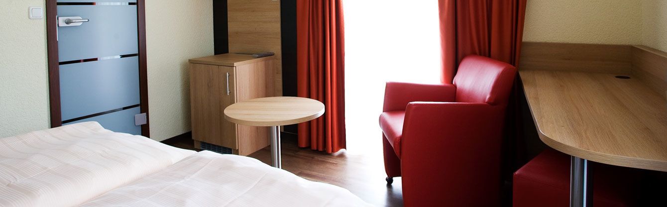 Een compacte hotelkamer met een bed, een klein tafeltje, een rode stoel, een houten kast en een inloopkast.