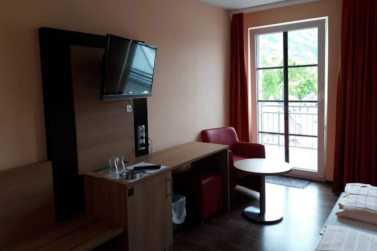 A hotel room with a TV mounted on the wall, a small desk with a chair, a round table, and a bed with neatly folded towels.