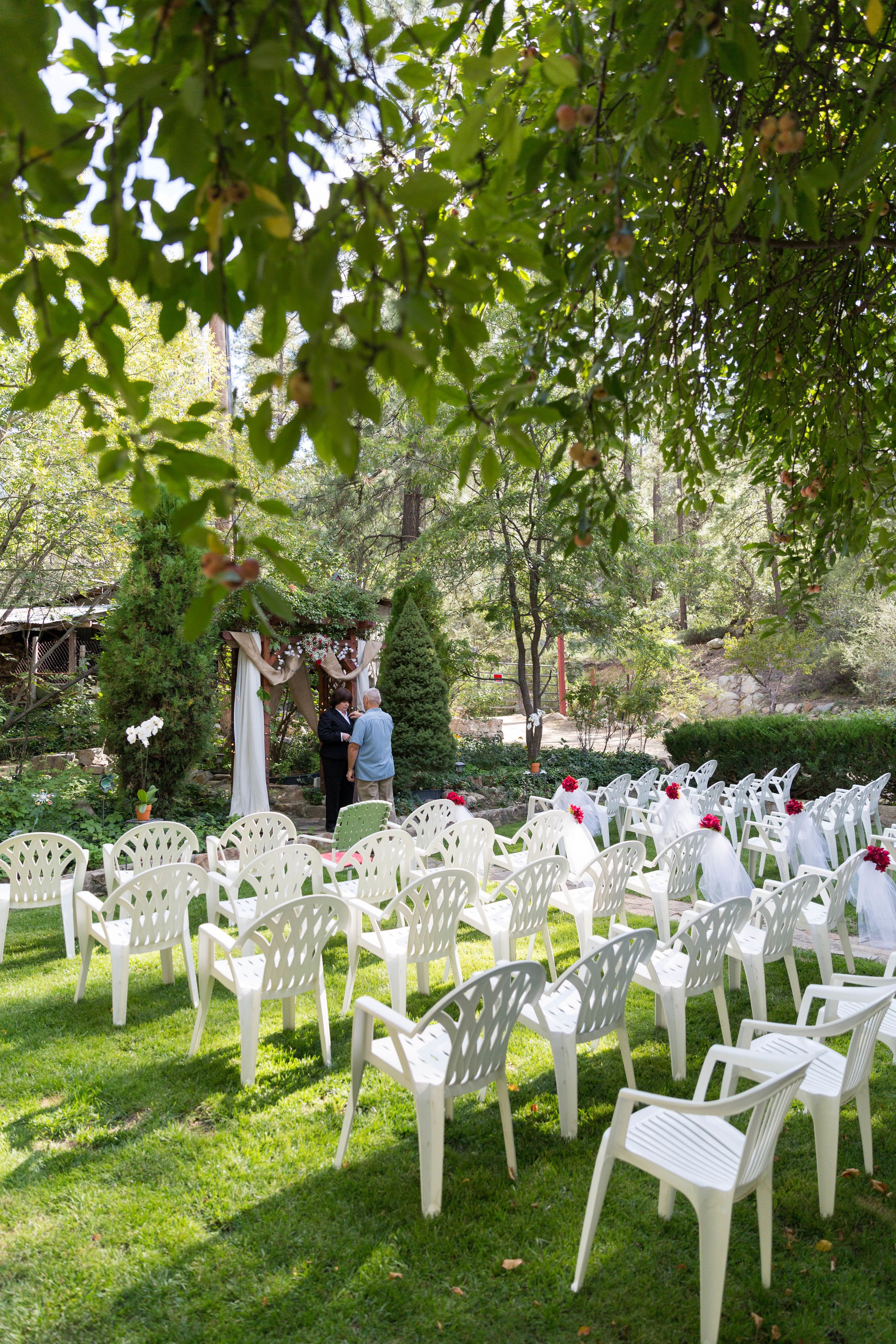 Un salón de bodas al aire libre con sillas de plástico blanco dispuestas en filas, un pequeño altar con tela blanca y follaje al fondo.