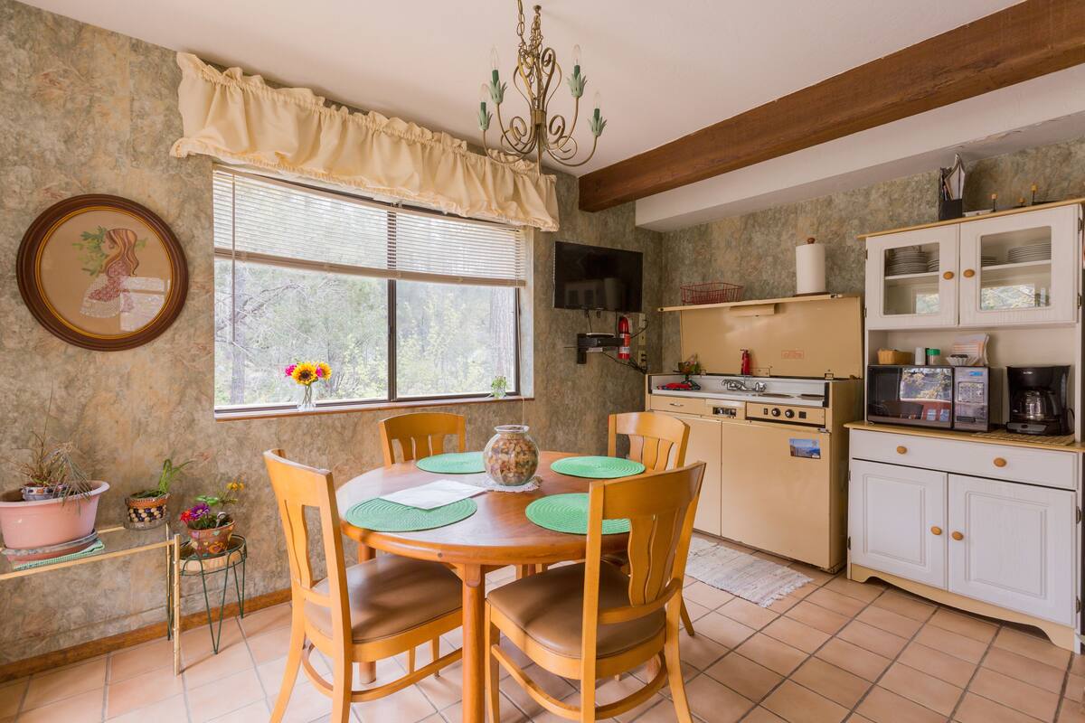 Una cocina con sillas de madera, una mesa de comedor, una araña, una ventana con persianas y un microondas.