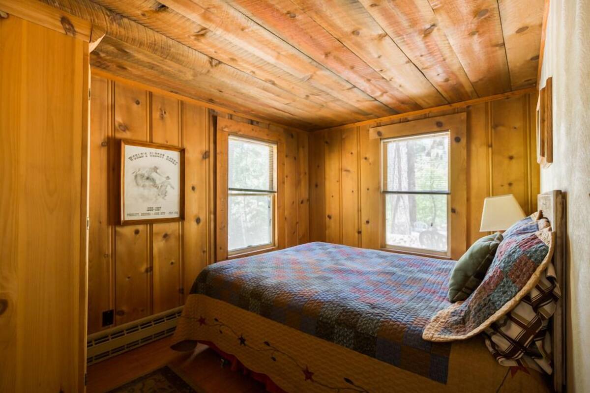 Un dormitorio de cabaña de madera con colcha, dos ventanas, un cuadro enmarcado y una silla con asiento acolchado.
