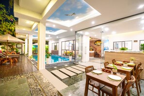 A modern indoor pool area with a glass ceiling, wooden deck, and a dining area with tables set for guests. - Hoi An Grace River Villa (Da Nang)