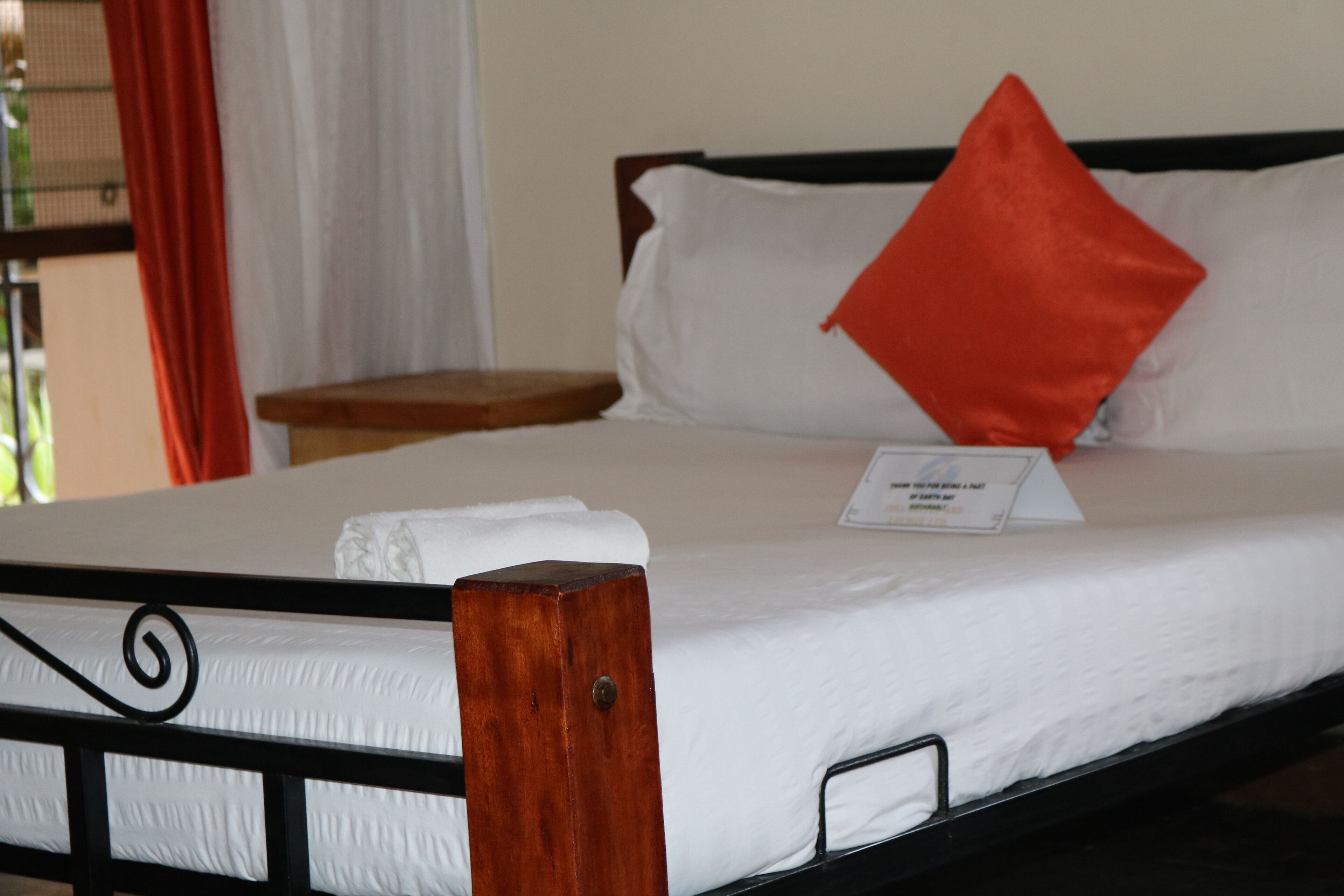 A neatly made bed with white bedding and a single red pillow, a wooden headboard, and a small bedside table with a lamp.