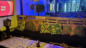 A cozy seating area with a wooden bench and colorful cushions, a table with a patterned tablecloth, and a wall-mounted TV displaying "THIS MAY". - Tisa Suites & Lounge (Kisumu)