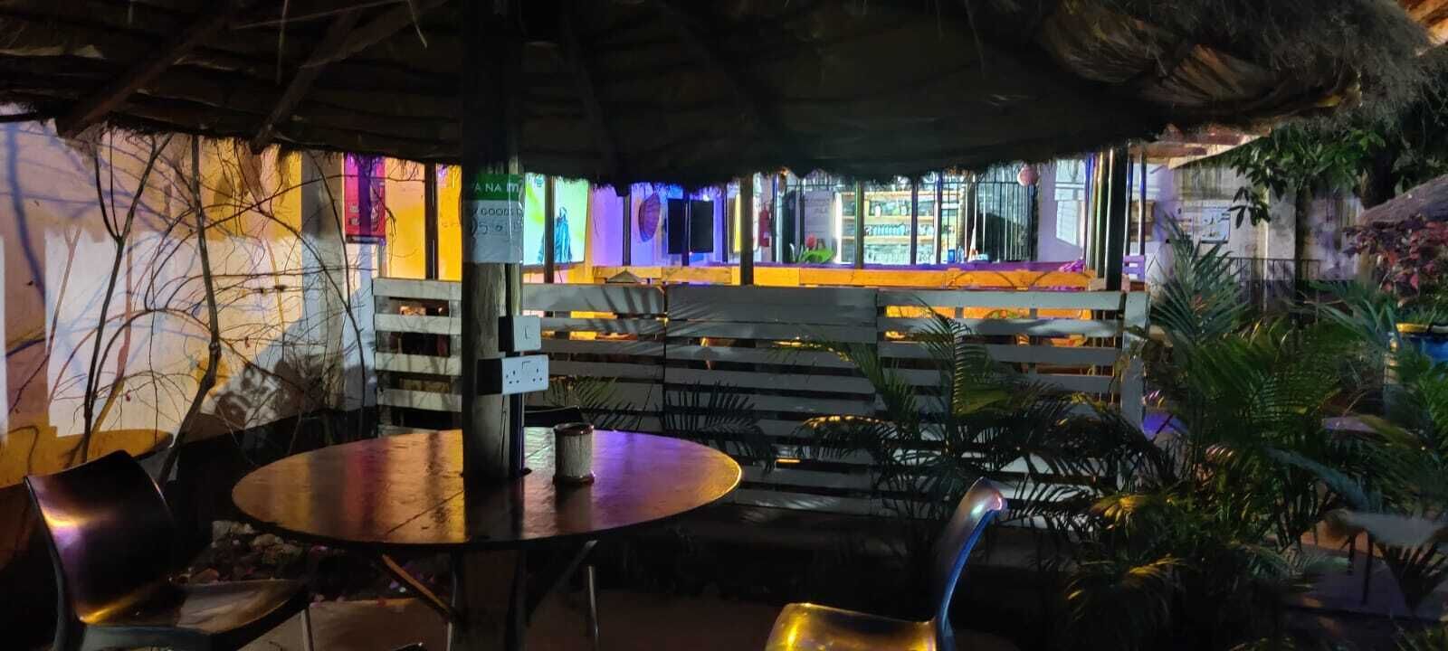An outdoor seating area with tables and chairs, a thatched roof, and a view of a colorful interior space.