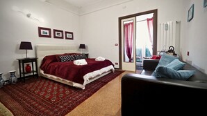 Ett sovrum med en säng, en soffa, en röd, mönstrad matta, två sängbord med lampor och ett fönster med gardiner.