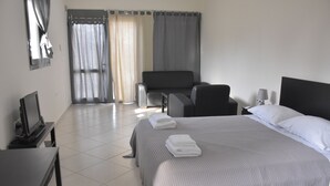Habitación de hotel con cama, escritorio con televisor, una silla, mesita de noche con lámpara y ventana con cortinas.