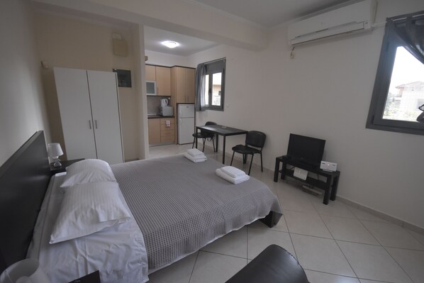 A hotel room with a bed, a desk, a chair, a television, and a kitchenette in the background.