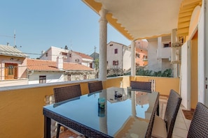 Varanda com mesa de vidro e cadeiras de vime, com vista para uma área residencial com telhados de terracota.