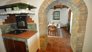 Une cuisine équipée d’une cuisinière, d’un four et d’étagères remplies de vaisselle. On y trouve également un coin repas avec une table et des chaises, ainsi qu’une arche en pierre qui mène à un escalier.