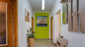 Un couloir avec une porte en bois, un panier en osier contenant une plante, un tableau représentant un voilier et un mur blanc orné d’un miroir et de crochets.