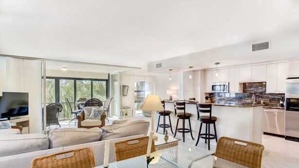 A modern living space with a glass dining table, wicker chairs, a sofa, and a kitchen area with a bar. - Kings Crown 210 (Sanibel)