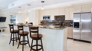 A modern kitchen with a central island, bar stools, stainless steel appliances, and a flat-screen TV mounted on the wall. - Kings Crown 210 (Sanibel)