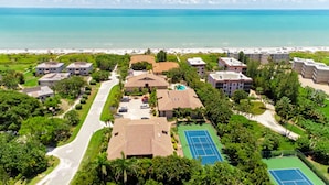 An aerial view of a coastal residential area with multiple buildings, a tennis court, and a beach in the background. - Coquina 1F (Sanibel)