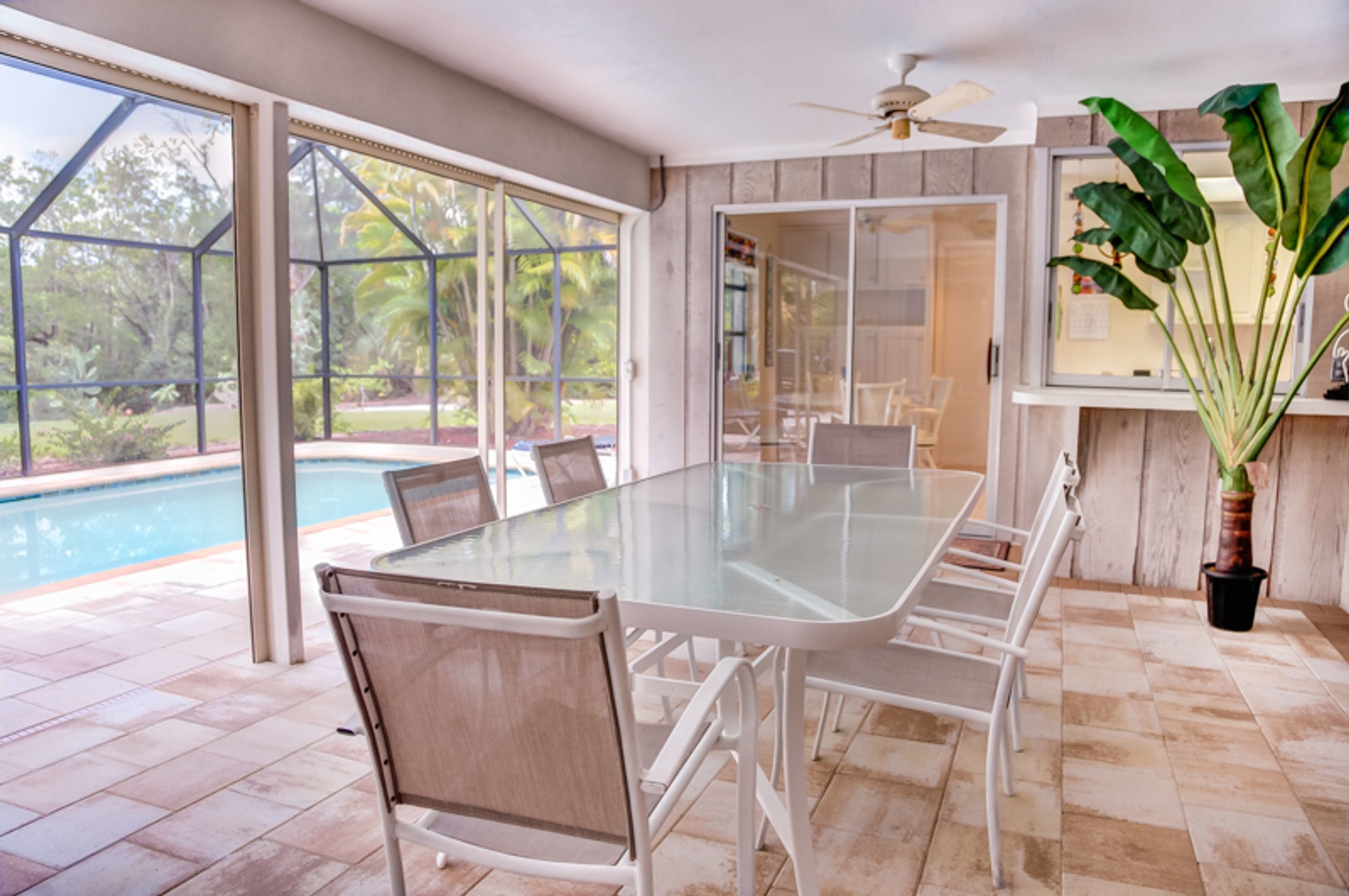 A spacious indoor area with a glass table and chairs, large windows, a ceiling fan, and a view of a swimming pool.