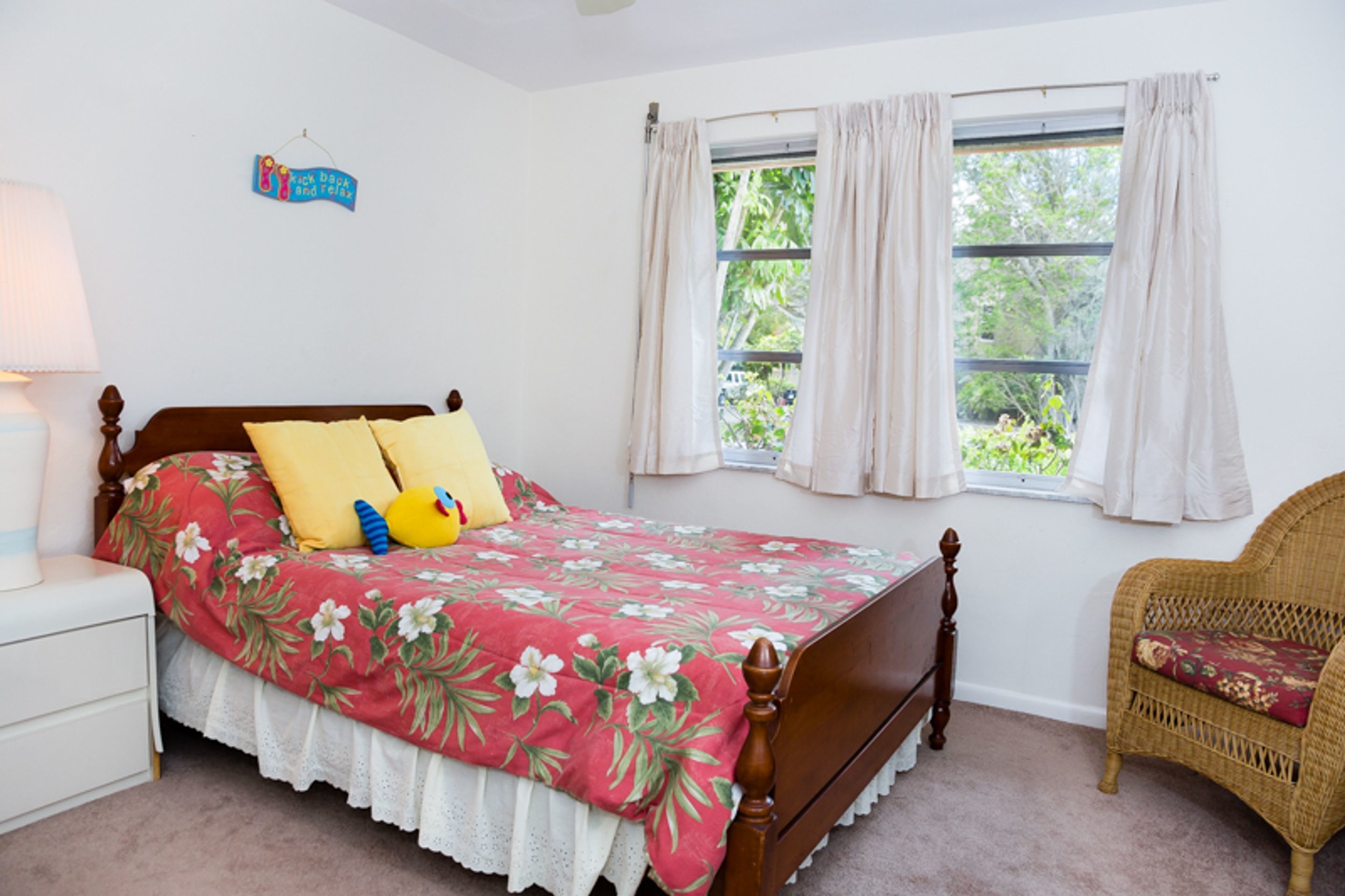 Ein Schlafzimmer mit einem hölzernen Bett, einem Nachttisch, einem Korbsessel und einem Fenster mit Vorhängen.