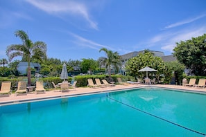A swimming pool with lounge chairs and umbrellas, surrounded by palm trees and a building in the background. - Mariner Point 1083 (Sanibel)