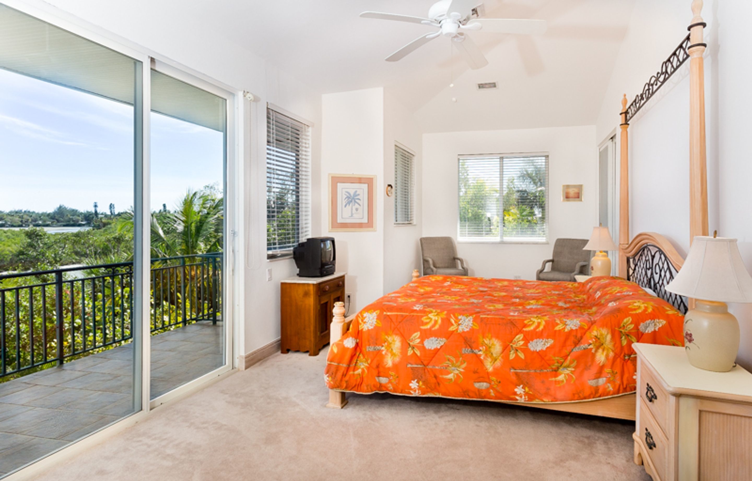 Une chambre à coucher comprenant un lit, une table de chevet, une chaise, un bureau, un ventilateur de plafond et un balcon avec vue sur la verdure.