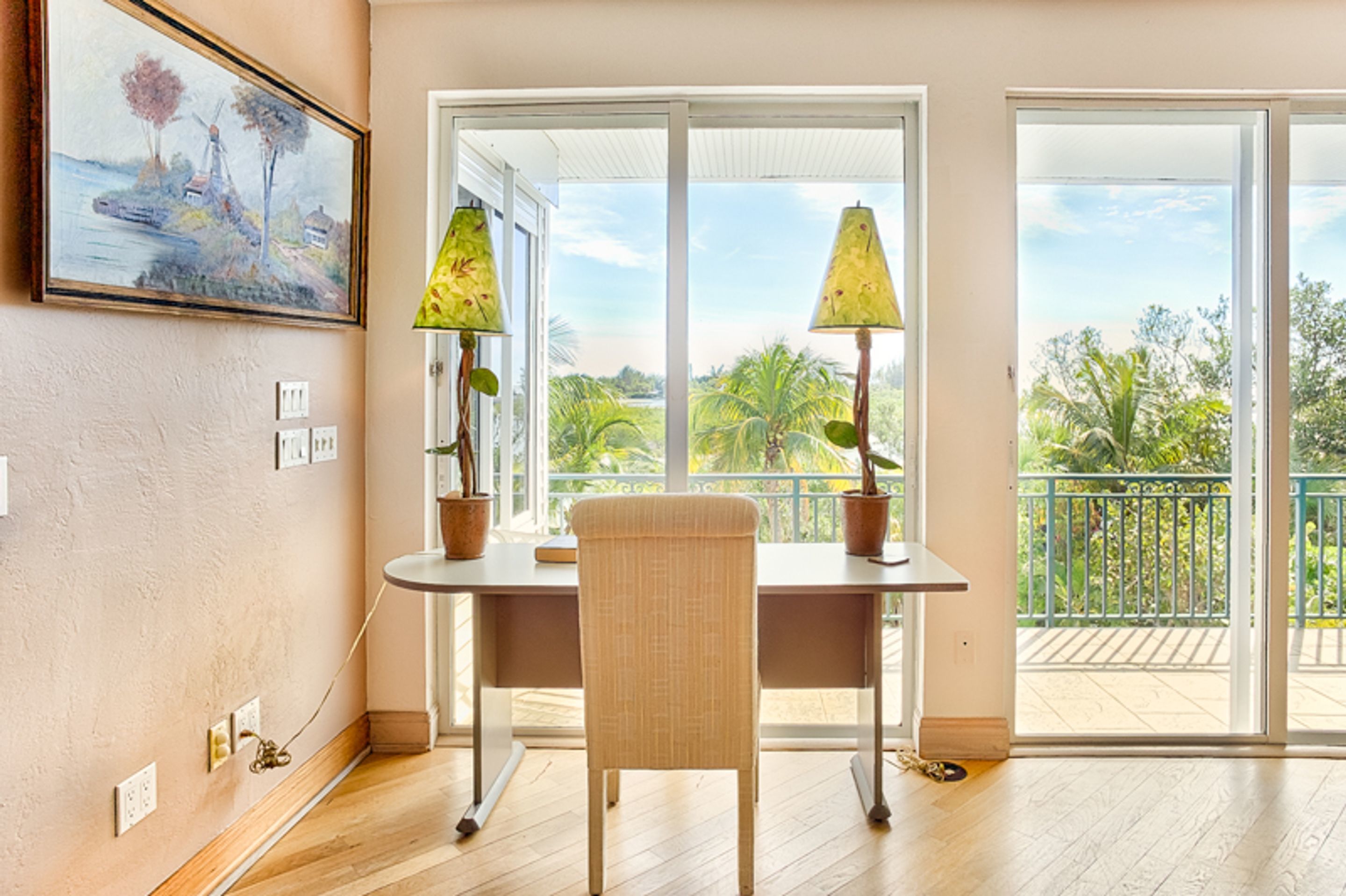 A room with a wooden floor, a table with two lamps, a chair, a painting on the wall, and a balcony with a view of palm trees.