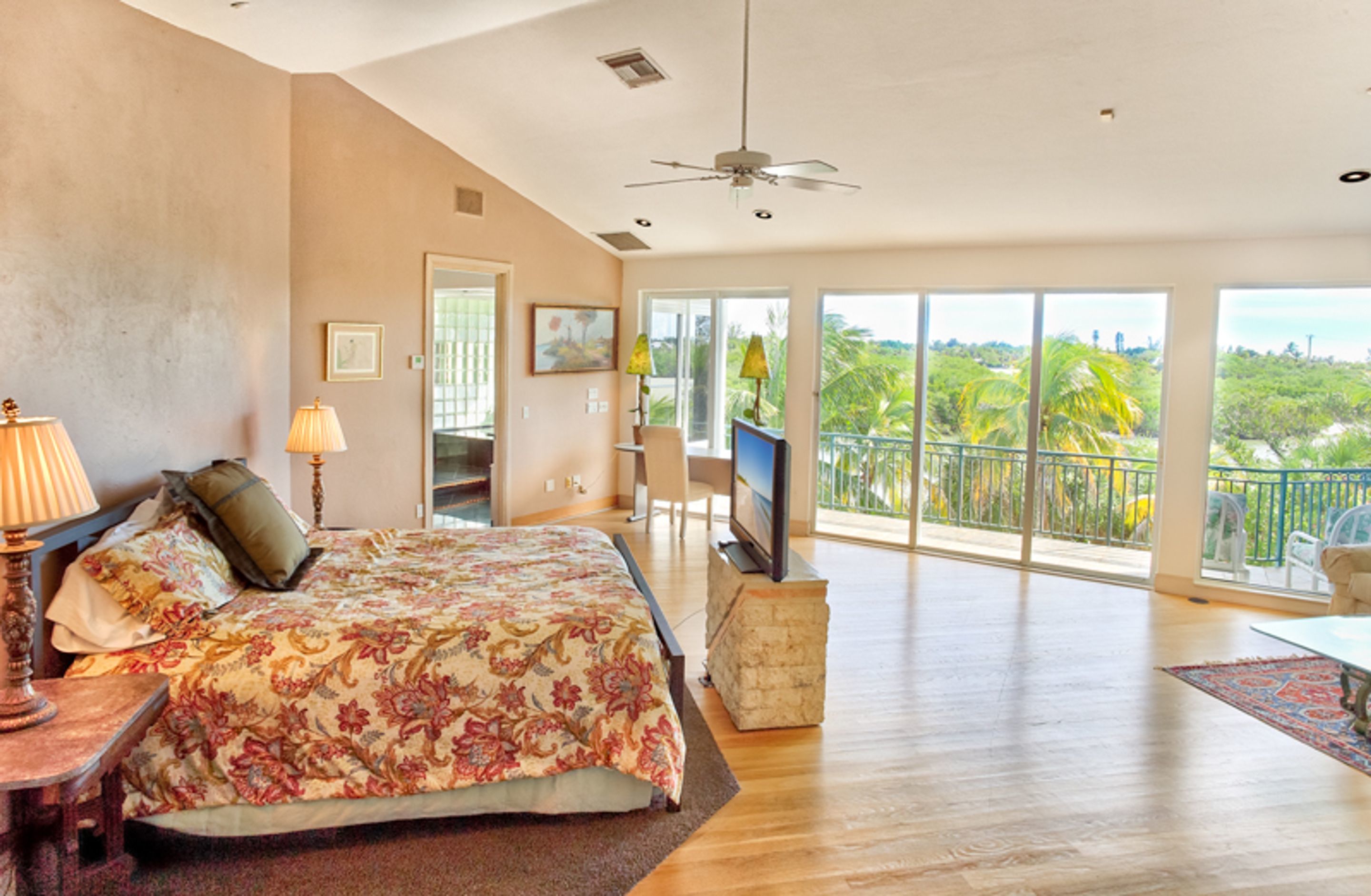A spacious bedroom with a large bed, a TV, and a balcony with a view of greenery.