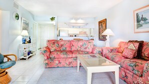 Een woonkamer met een bloemenbank, een glazen salontafel en een keuken op de achtergrond.