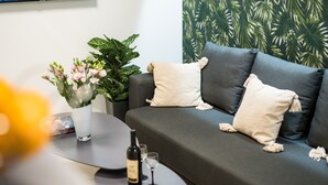 Un saló modern amb un sofà gris, dos coixins blancs, una taula petita amb una ampolla i gots, i una planta en test amb flors roses.