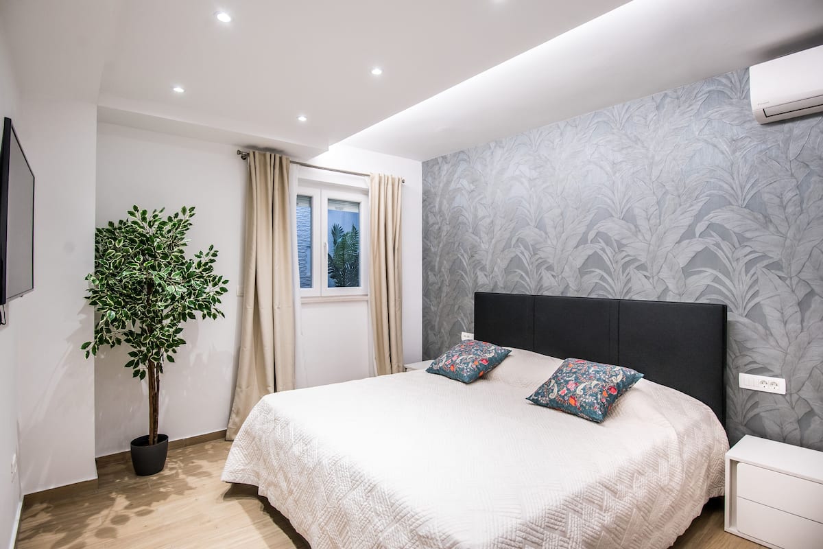 A modern bedroom with a large bed, a potted plant, a window with curtains, and a wall-mounted air conditioner.