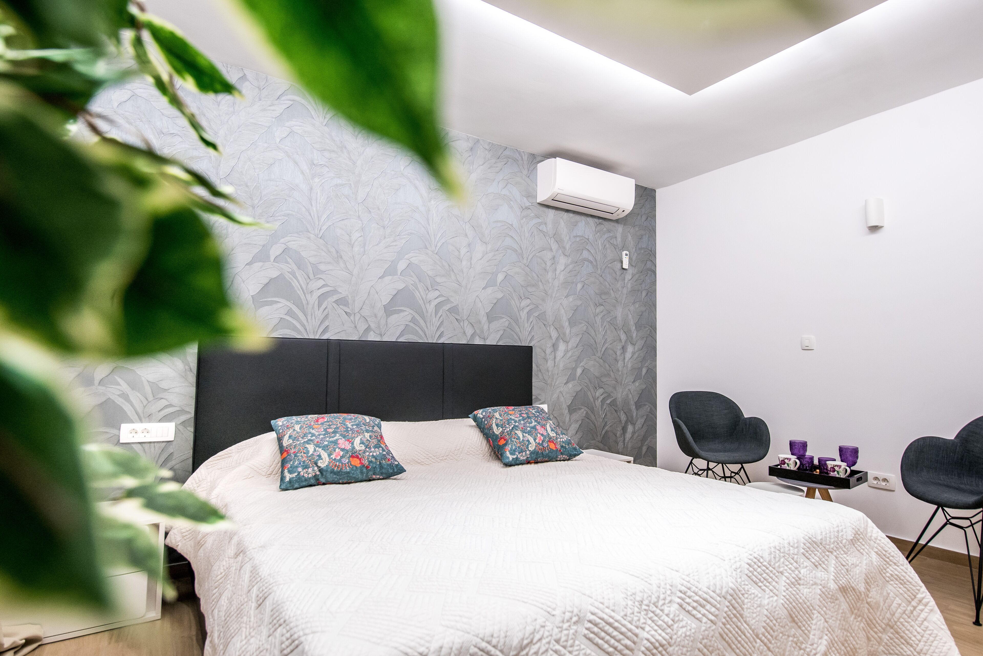 A modern bedroom with a large bed, floral-patterned pillows, a dark headboard, and a side table with purple cups.