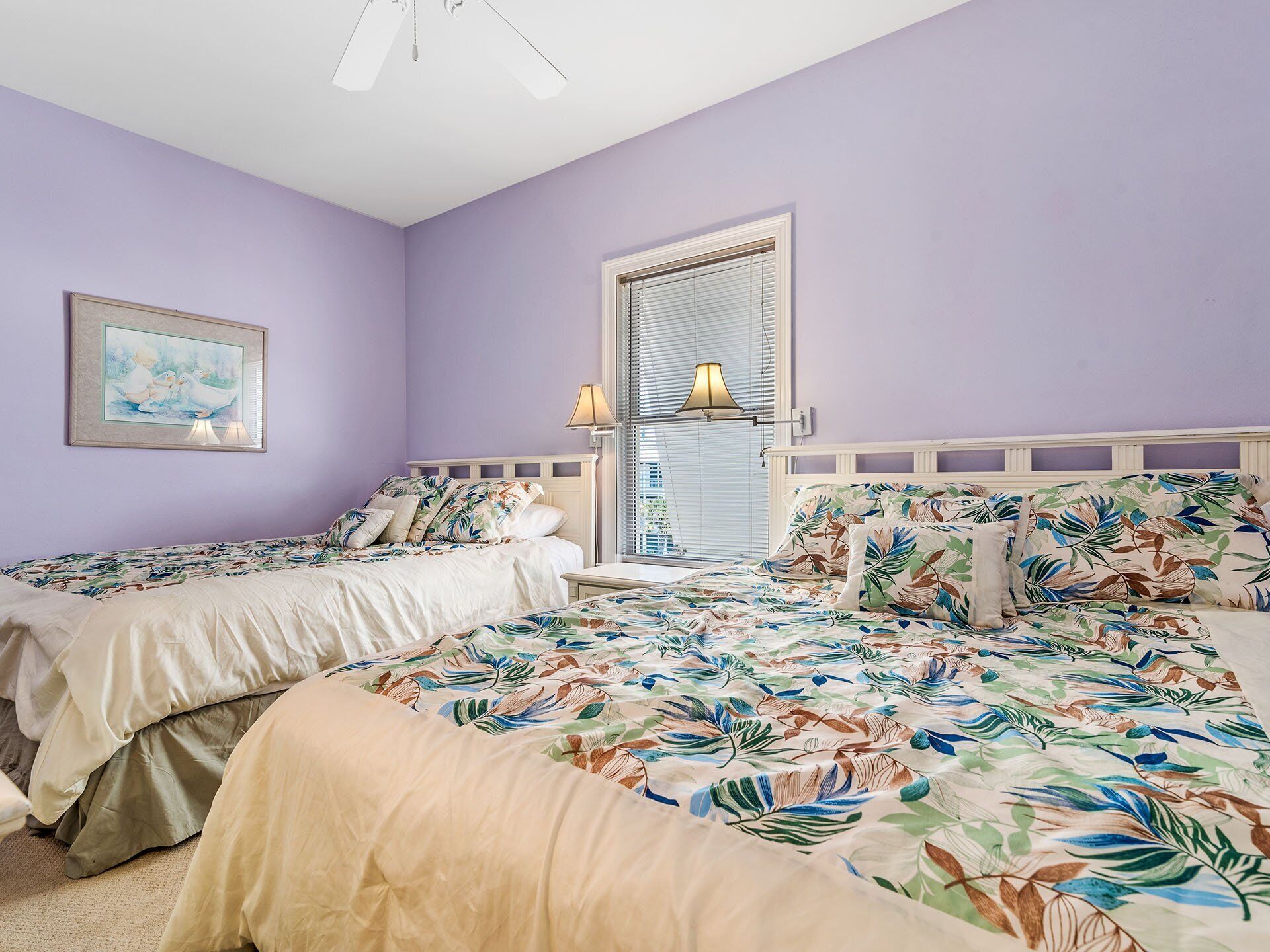 A bedroom with two single beds, a large bed with a floral bedspread, a ceiling fan, a window with blinds, and a framed picture on the wall.