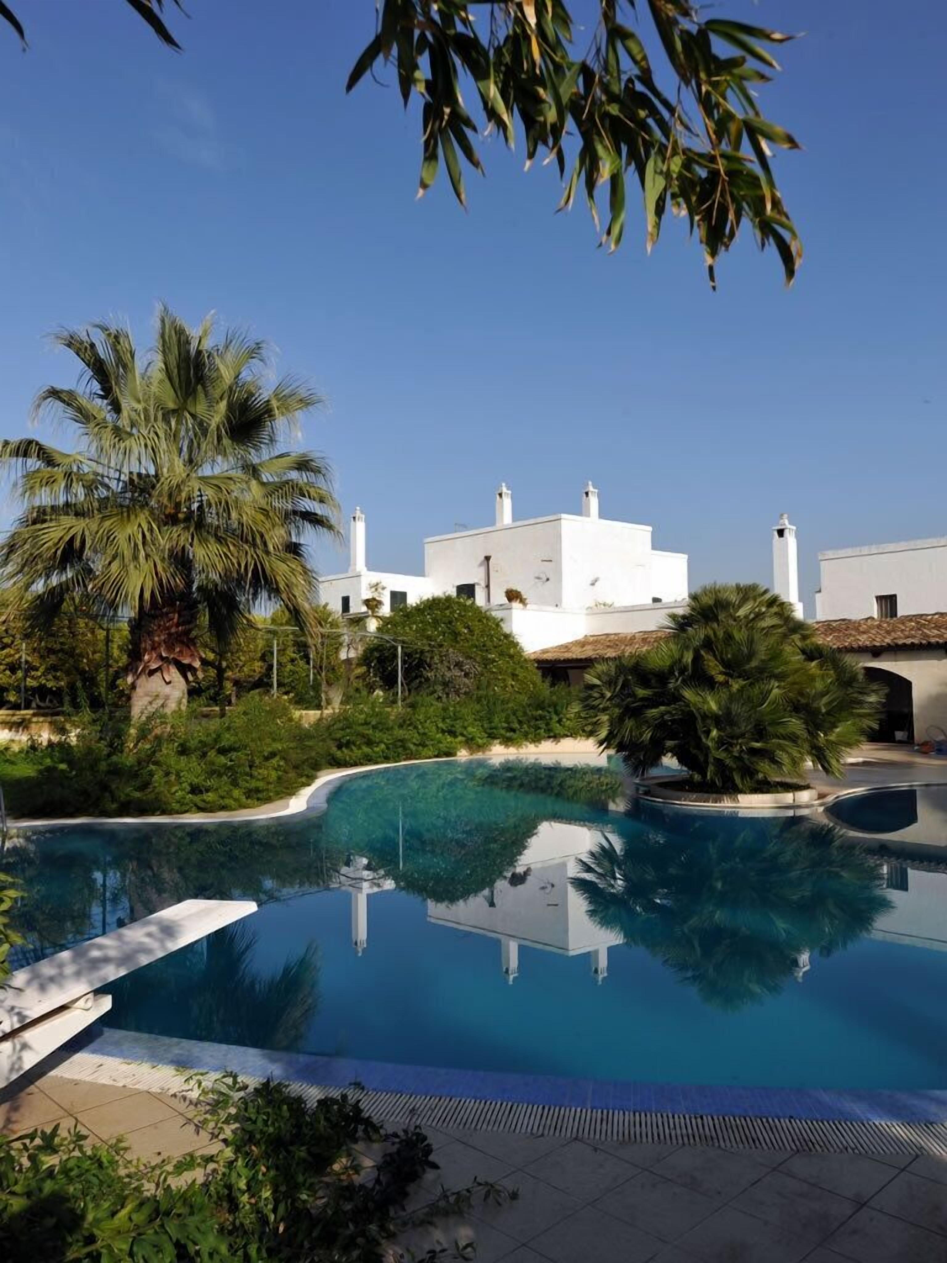 A swimming pool with a clear blue water, surrounded by greenery and white buildings in the background.