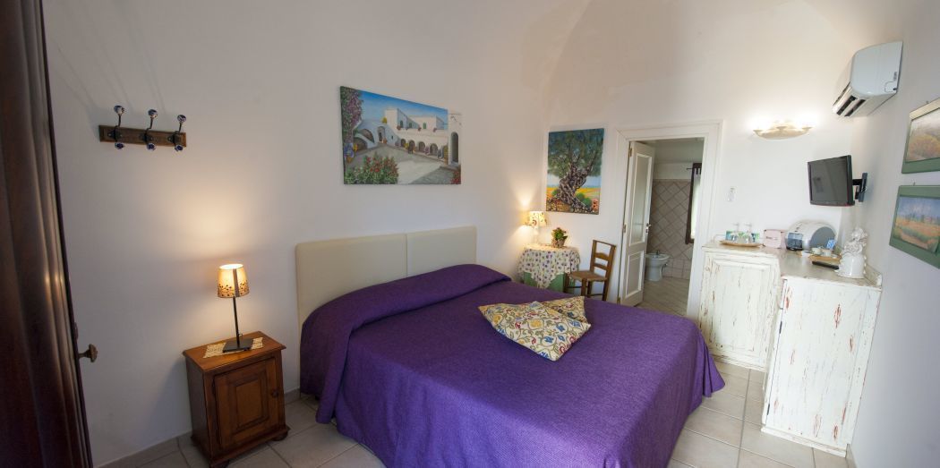 Ein Schlafzimmer mit einem Bett, einem Nachttisch, einer Lampe und einem kleinen Tisch mit einem Stuhl. Eine Tür führt in einen anderen Raum, und eine Wandklimaanlage ist angebracht.