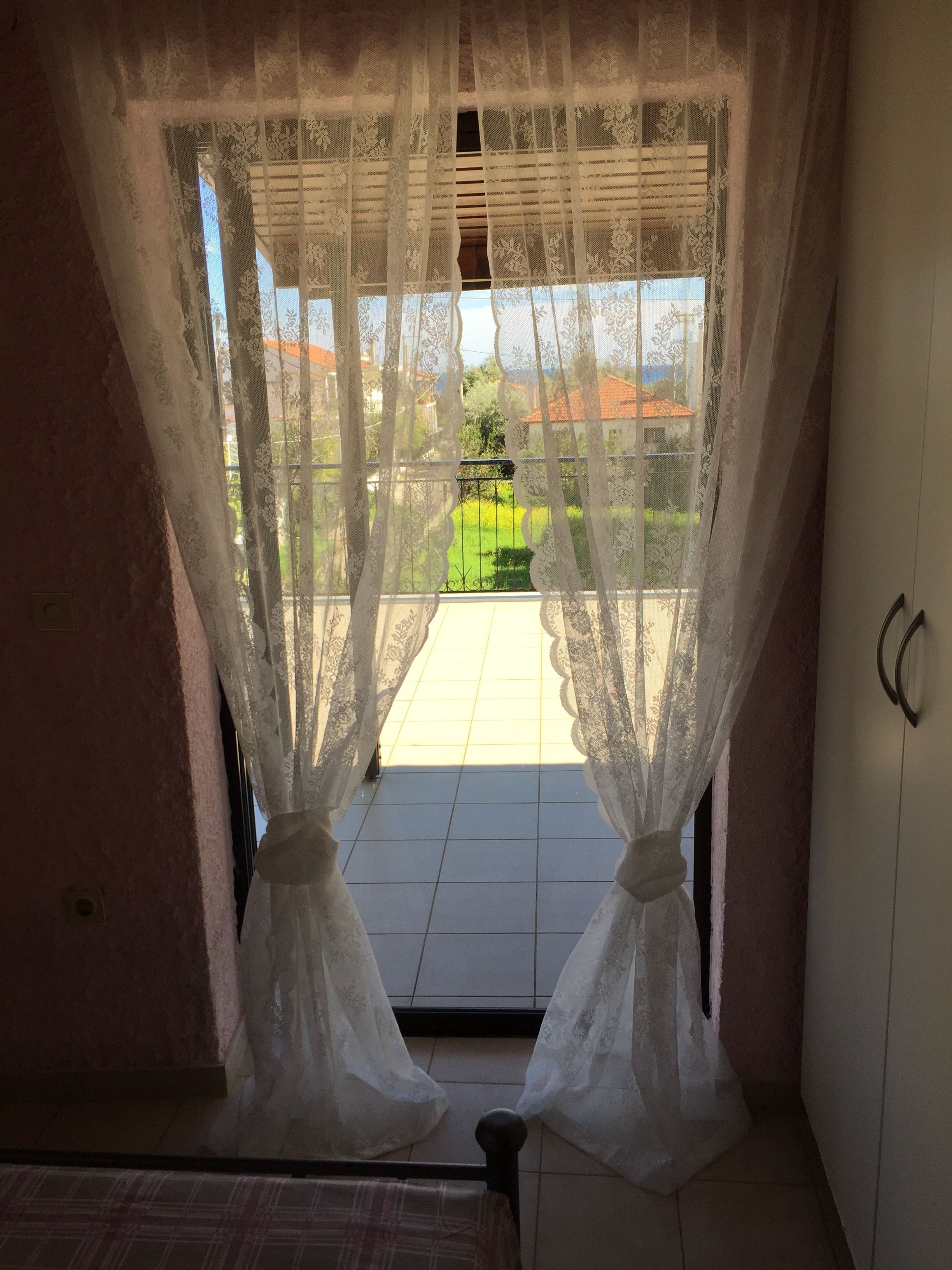 A room with a view through lace curtains to a balcony with a tiled floor and a green lawn.
